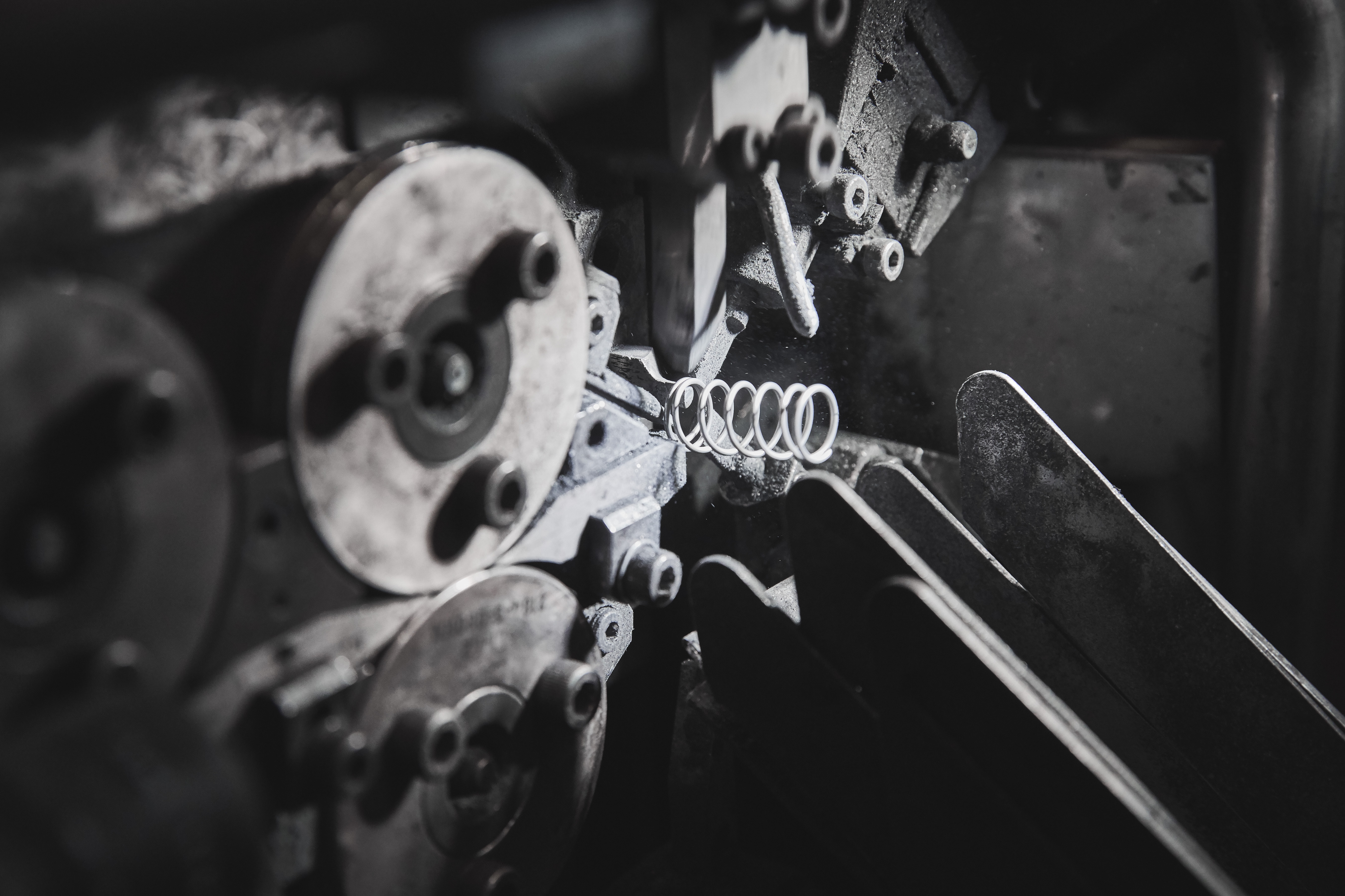 Close-up of a metal coil spring being formed inside a spring coiling machine, with wire feeding through rollers under low-light conditions.