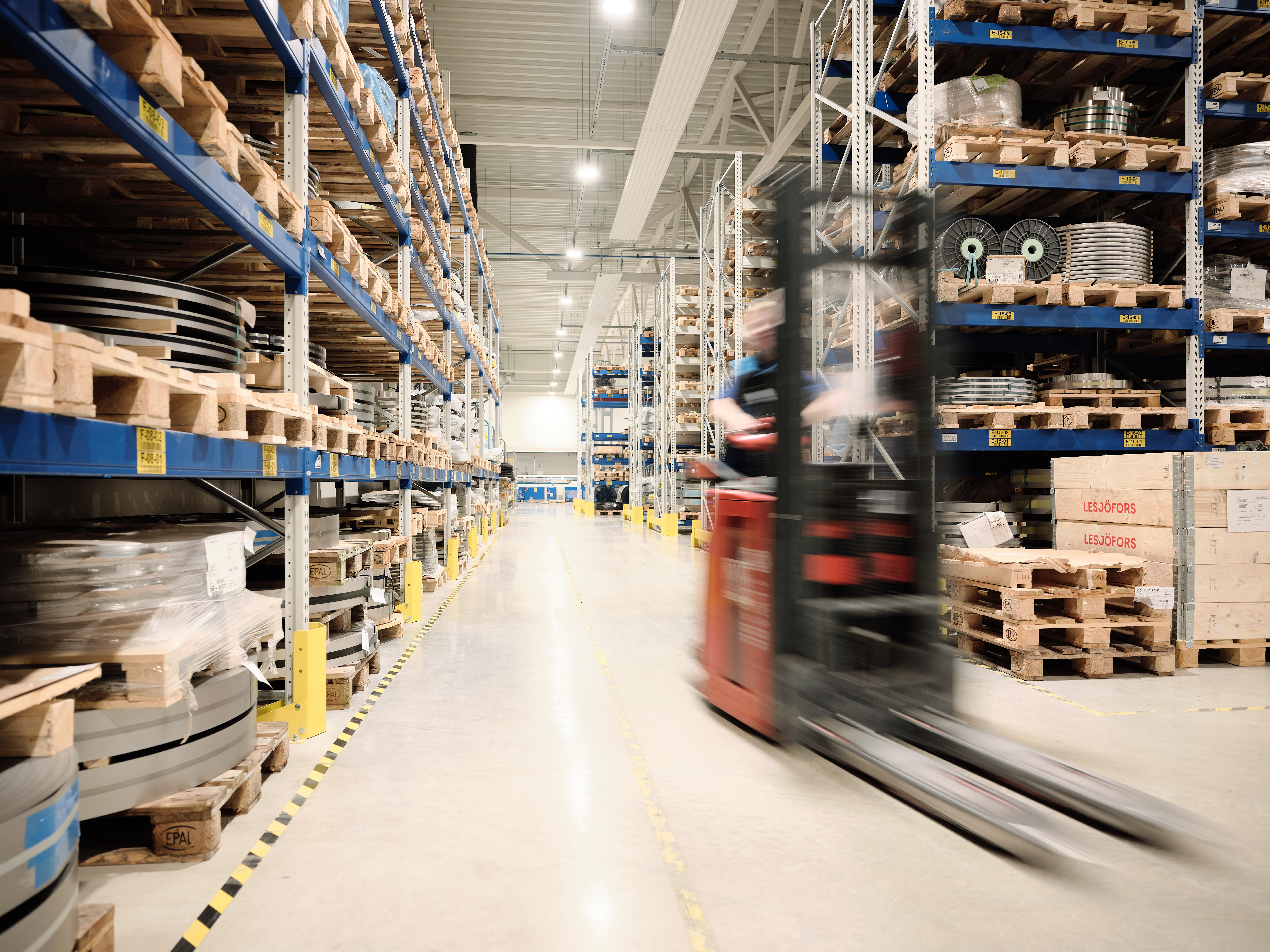 A motion-blurred forklift drives through a wide warehouse aisle, with tall racks on both sides holding pallets of metal materials and packaged goods.